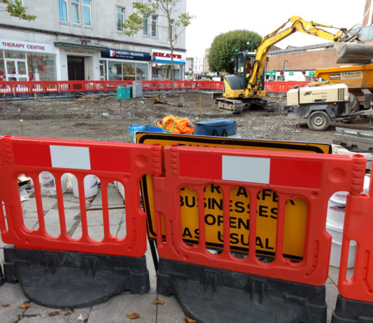 A sign saying businesses open as usual obscured by a roadworks barrier.