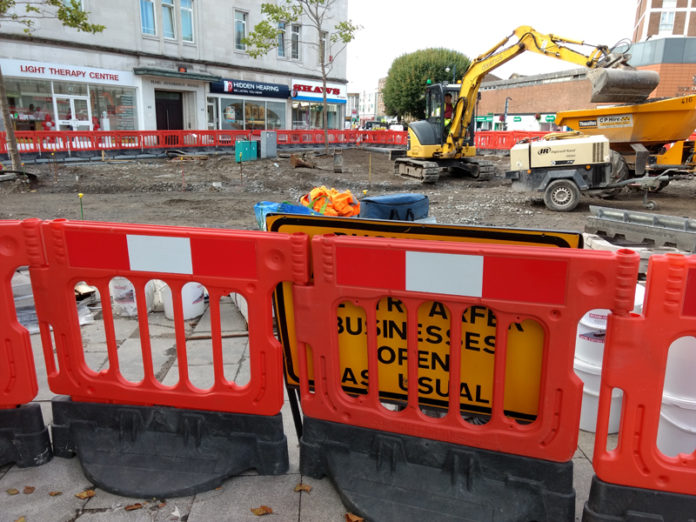 A sign saying businesses open as usual obscured by a roadworks barrier.